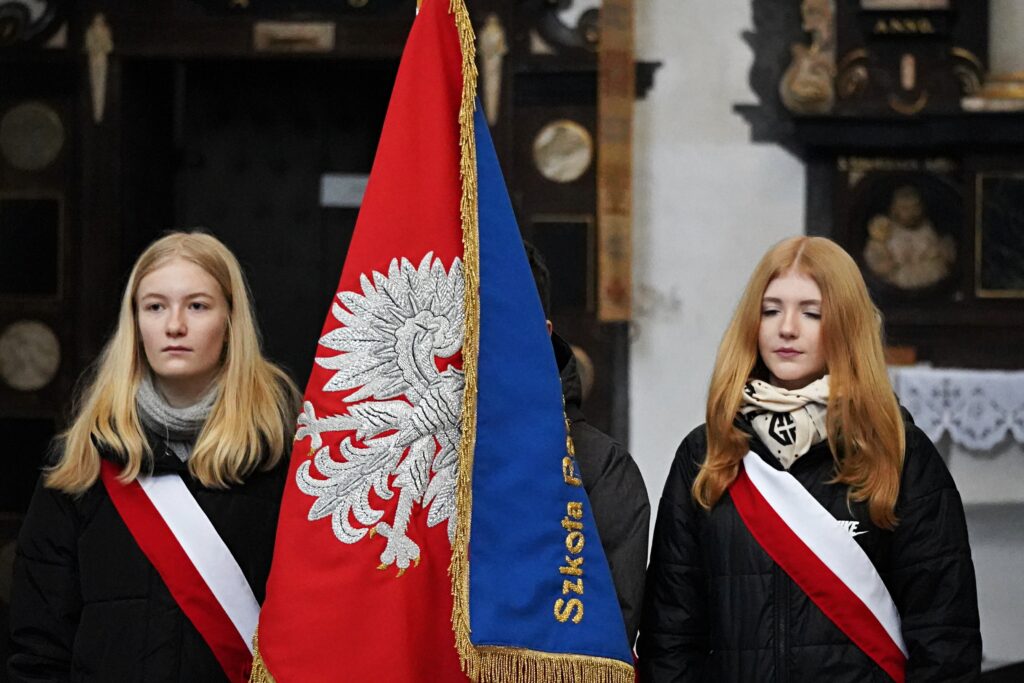 "Dwie dziewczyny w strojach galowych z biało-czerwonymi szarfami trzymają sztandar szkoły. W tle widoczne są elementy kościelnego wyposażenia."