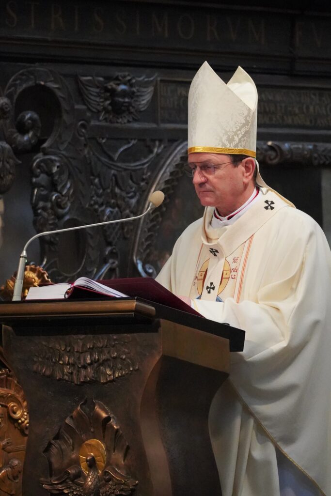 "Zbliżenie na biskupa przy ambonie podczas homilii. Ma na sobie białą mitrę oraz zdobioną szatę liturgiczną. W tle widoczne są rzeźbione elementy ołtarza."