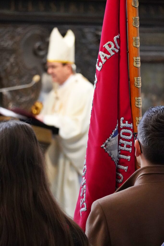 Fragment uroczystości w kościele. Na pierwszym planie widoczny czerwony sztandar, za którym znajduje się biskup przemawiający podczas mszy."