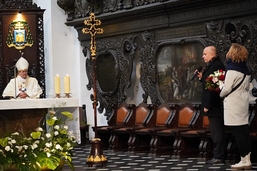 Biskup siedzi obok ołtarza, natomiast mężczyzna z mikrofonem i kobieta z kwiatami przemawiają przy bogato zdobionym obrazie w tle.