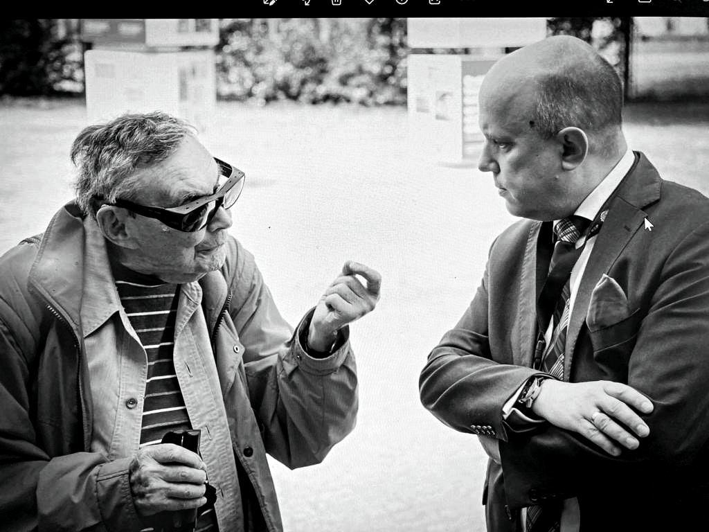 The black-and-white photograph captures a conversation between two men. On the left is Marian Turski, a Holocaust survivor, historian, and journalist, dressed in a light jacket and a striped shirt. He wears his distinctive thick-framed glasses and holds a cane in his hand. His facial expression is serious, and his hand gestures emphasize the importance of his words.

On the right stands Piotr Tarnowski, director of the Stutthof Museum. He is dressed in an elegant suit, tie, and pocket square. With his arms crossed, he listens attentively to his interlocutor, displaying focus and respect.

The photograph captures a moment of profound discussion, likely about the memory of Holocaust victims and the role of museums in preserving history. In the background, blurred elements of an outdoor space suggest a historical site or an open-air exhibition.