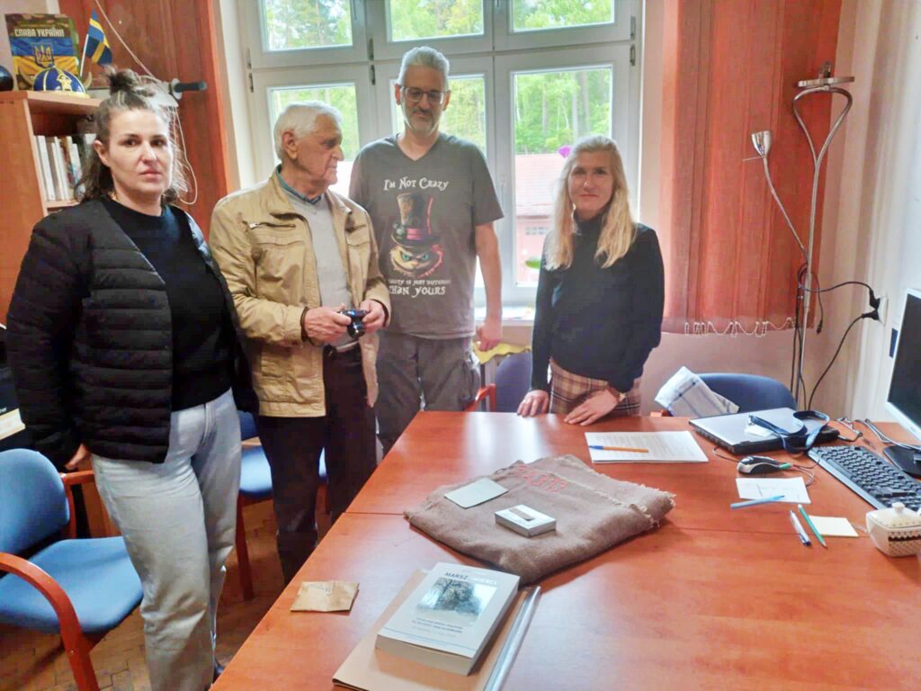 Cztery osoby, w tym Pan Andrzej Przyklęk, stoją przy biurku w pomieszczeniu muzealnym. Na blacie leżą przekazane pamiątki po więźniach KL Stutthof: koc, naszywka, papierośnica oraz dokumenty.