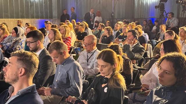 Publiczność zgromadzona na konferencji. Widoczne są dziesiątki osób siedzących w rzędach i słuchających panelu. Wśród nich osoby noszące słuchawki i wykonujące notatki. W tle metalowa hala i światła oświetleniowe.