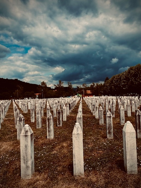 Widok na cmentarz ofiar ludobójstwa w Srebrenicy. Setki białych nagrobków ustawionych w równych rzędach ciągną się aż po horyzont. Nad cmentarzem gromadzą się ciężkie, burzowe chmury.