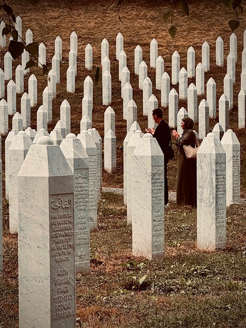 Dwie osoby stoją wśród rzędów białych nagrobków na cmentarzu w Srebrenicy. Mężczyzna i kobieta w chustach modlą się, w tle widać wzgórze i dalsze rzędy grobów. Na pierwszym planie widoczne inskrypcje na kamieniach nagrobnych w języku arabskim i łacińskim.