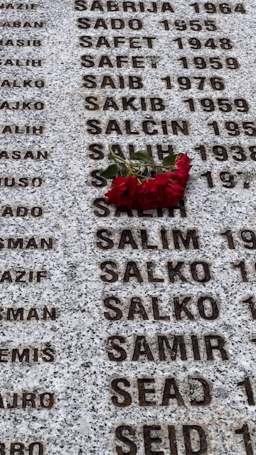 Fragment marmurowej tablicy pamiątkowej z wyrytymi imionami ofiar. Na nazwiskach ułożone są czerwone róże – symbol pamięci i żałoby.