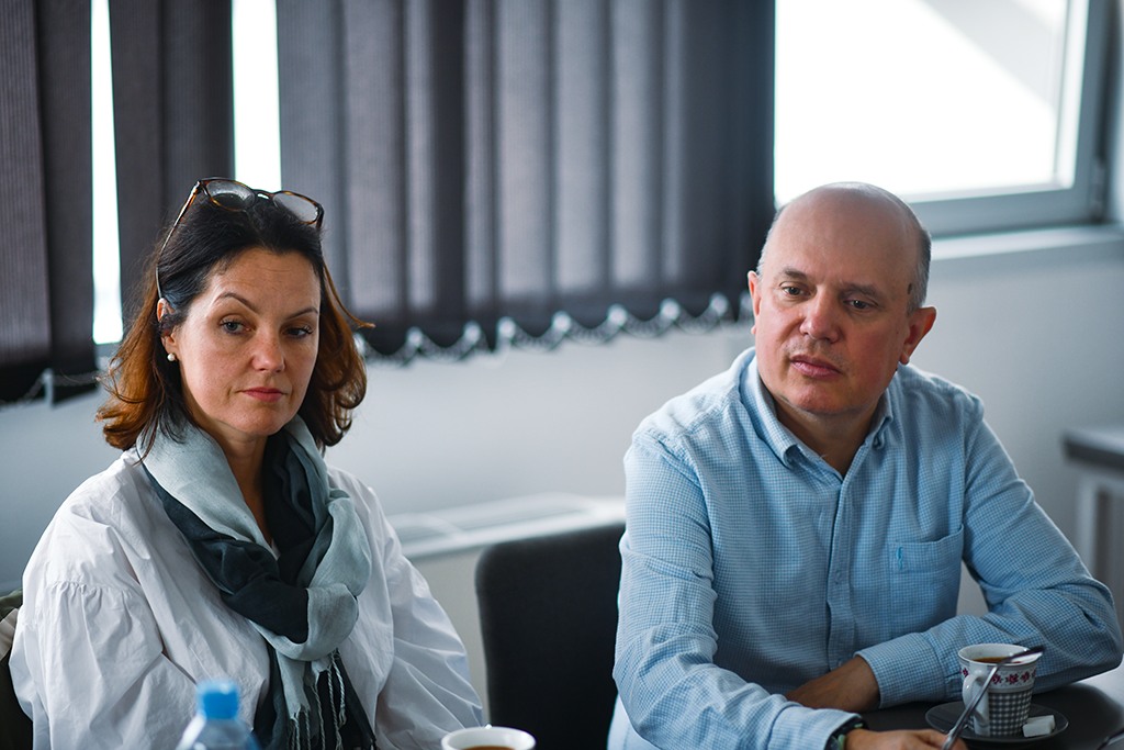 Kobieta i mężczyzna siedzą przy stole podczas spotkania. Kobieta ubrana w białą koszulę i szalik, mężczyzna w niebieskiej koszuli. Przed nimi filiżanki i butelka wody. W tle zasłonięte okna i neutralne wnętrze sali spotkań.