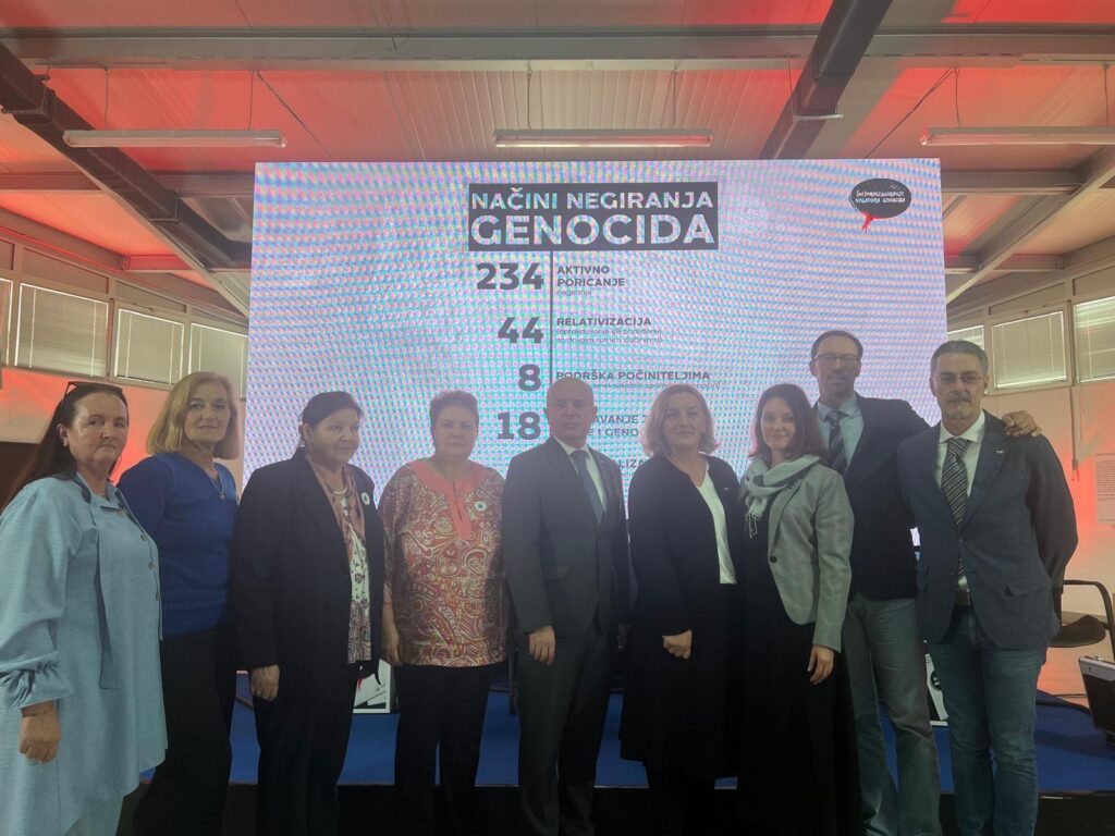 Grupa dziesięciu osób pozuje do wspólnego zdjęcia na tle dużego ekranu z napisem w języku bośniackim „Načini negiranja genocida” (Sposoby negowania ludobójstwa). Wśród grupy znajduje się dwoje pracowników Muzeum Stutthof – kobieta w jasnym szalu oraz mężczyzna w garniturze.