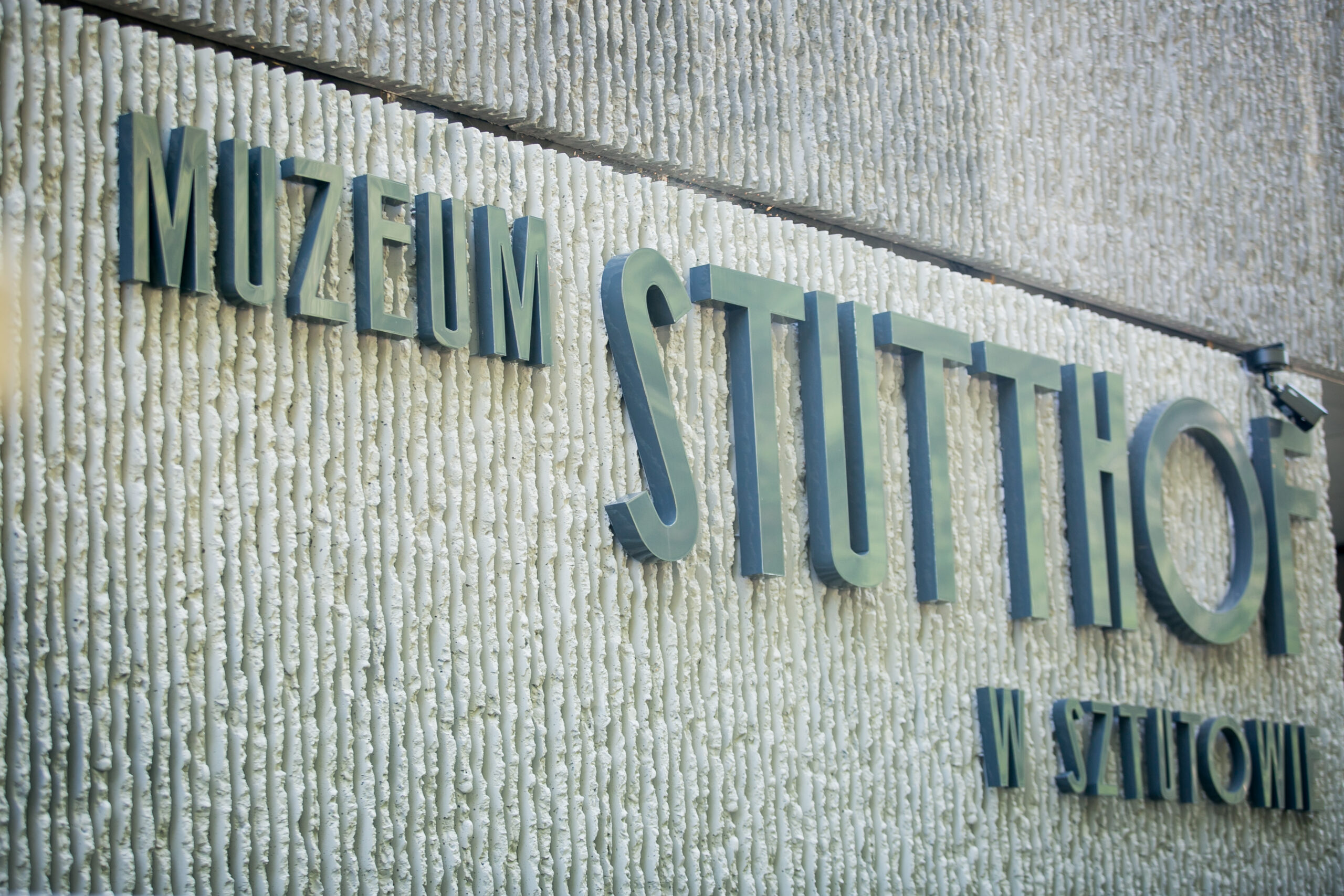 Zbliżenie na elewację budynku z dużym, metalowym napisem „Muzeum Stutthof w Sztutowie”. Litery kontrastują z jasną, fakturowaną powierzchnią ściany.
