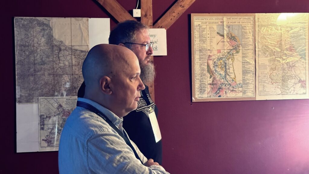 Dwóch uczestników seminarium stoi obok siebie, w tle widoczne stare mapy i plansze historyczne na czerwonej ścianie.