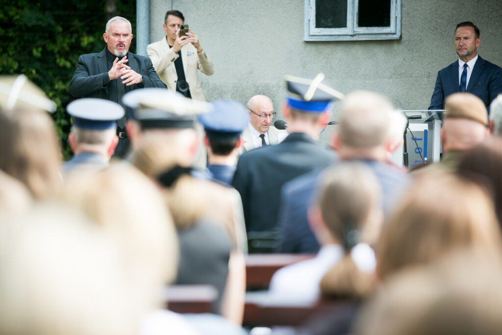 Ujęcie z perspektywy widowni; po lewej tłumacz PJM, w głębi przy mównicy przemawia prelegent, wokół mundury i goście uroczystości.