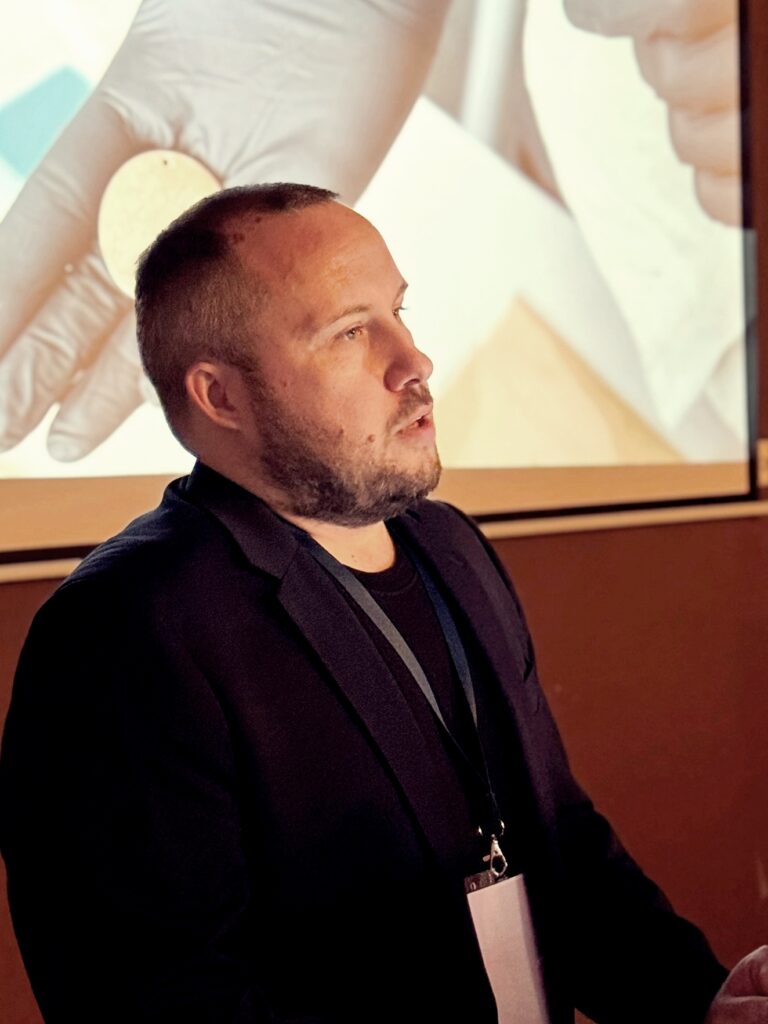Prelegent w ciemnej marynarce przemawia, za nim na ekranie wyświetlona jest prezentacja zbliżenia dłoni trzymającej monetę w rękawiczce ochronnej.