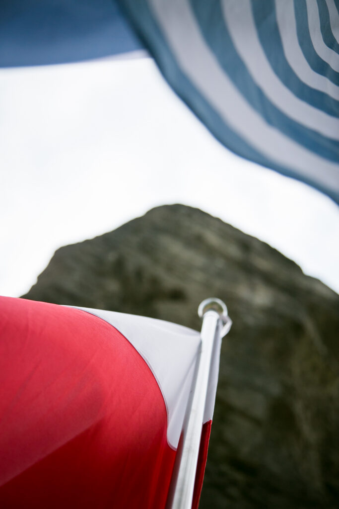 Zbliżenie na biało-czerwoną flagę Polski wznoszącą się ku górze obok fragmentu niebiesko-białego pasiaka obozowego, w tle monumentalny kamienny pomnik.