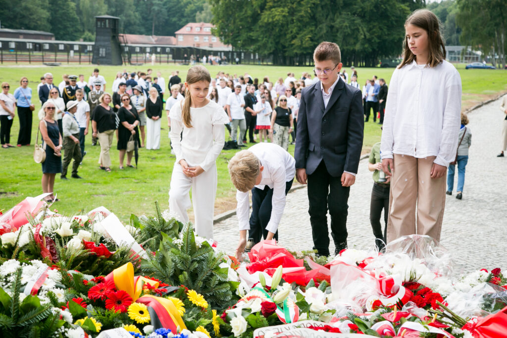 Grupa dzieci w białych i eleganckich strojach pochyla się nad miejscem pamięci, składając kwiaty i wieńce wśród licznie zgromadzonych uczestników uroczystości.