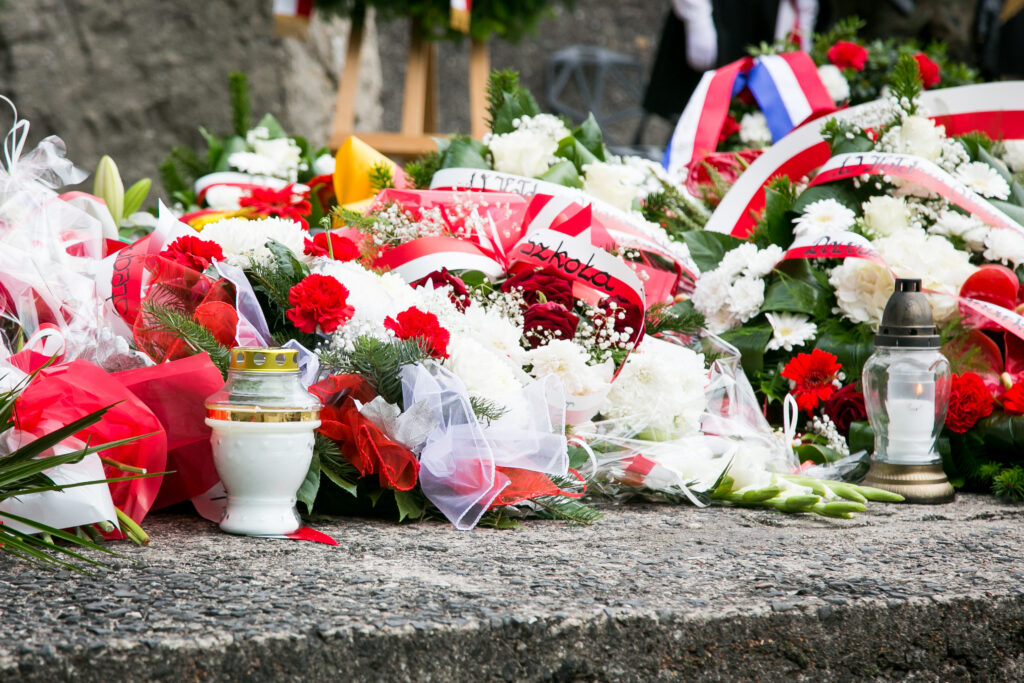 Zbliżenie na kolorowe wieńce, kwiaty i zapalone znicze ułożone pod pomnikiem – dominują czerwone i białe barwy, symbolizujące pamięć i hołd dla ofiar.