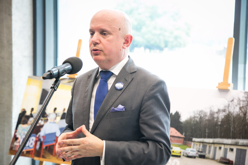 Dyrektor muzeum w szarym garniturze i niebieskim krawacie stoi przy mikrofonie i wygłasza przemówienie. Na klapie ma przypiętą rocznicową przypinkę, za nim widoczne plansze wystawowe.