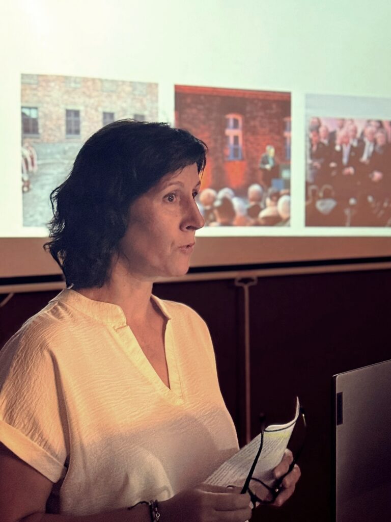 Kobieta w jasnej bluzce prowadzi wystąpienie, w tle ekran z obrazami przedstawiającymi sceny z wydarzeń w obozie Auschwitz.