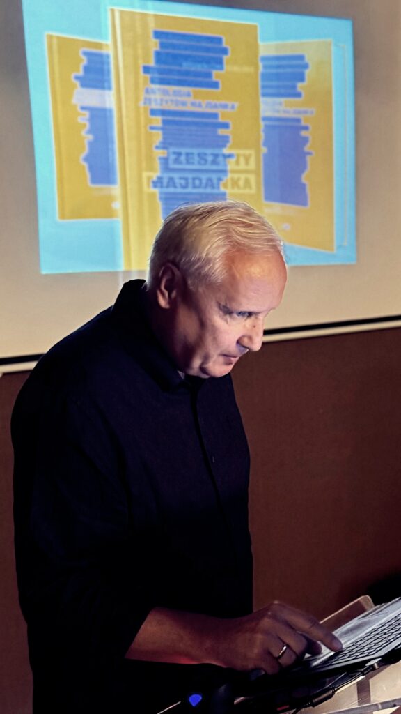 Mężczyzna w czarnej koszuli czyta notatki przy pulpicie, na ekranie za nim widoczna okładka publikacji „Zeszyty Majdanka”