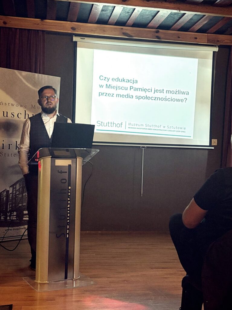„Mężczyzna w okularach, ubrany w białą koszulę i ciemną kamizelkę, stoi za mównicą z napisem ‘Stutthof’. W tle wyświetlona jest prezentacja multimedialna z pytaniem: ‘Czy edukacja w Miejscu Pamięci jest możliwa przez media społecznościowe?’. Obok widoczny fragment baneru Państwowego Muzeum Auschwitz-Birkenau. Sala ma drewniany sufit, uczestnicy siedzą na krzesłach, część osób zwrócona w stronę prelegenta.”