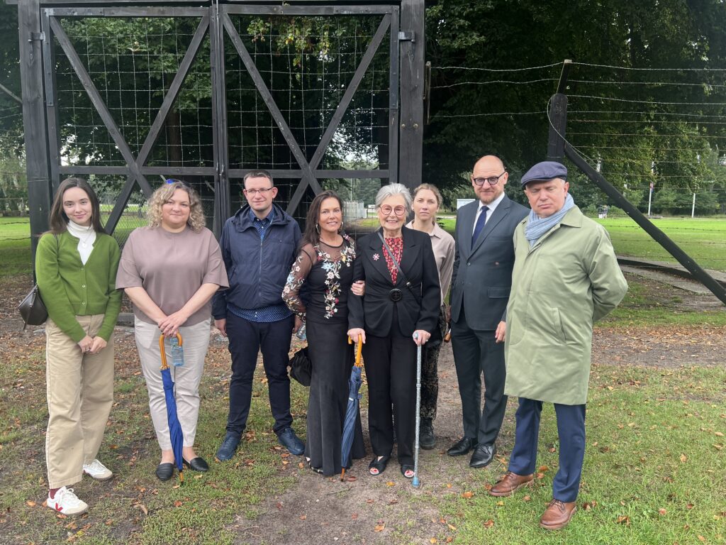 Grupa ośmiu osób stoi przed historyczną bramą obozu koncentracyjnego Stutthof, w tle widoczne fragmenty ogrodzenia z drutem kolczastym. W centrum grupy znajduje się starsza kobieta z laską, obok niej Ambasador Łotwy w Polsce Raimonds Jansons.
