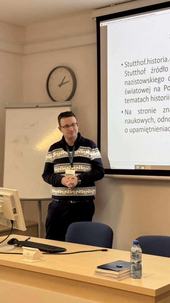 Mężczyzna w swetrze stoi obok ekranu z prezentacją i mówi do grupy; w tle widać tablicę suchościeralną i zegar