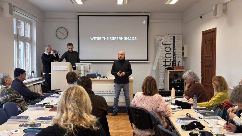 Prelegent stoi przed ekranem z dużym napisem ‘We’re the Superhumans’, a uczestnicy siedzą przed nim przy stołach i słuchają