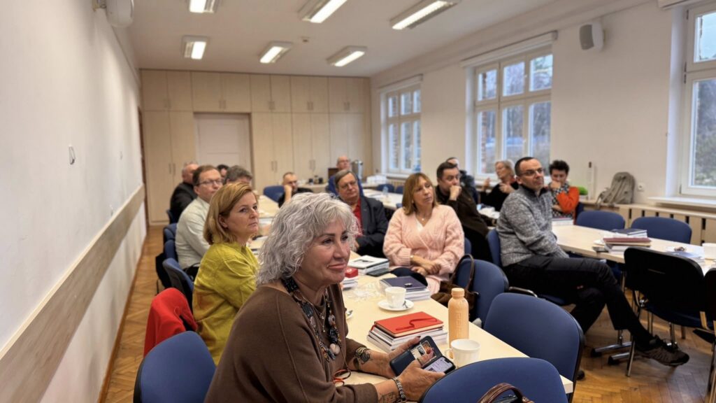 Grupa dorosłych uczestników siedzi przy stołach w jasnej sali szkoleniowej i słucha prowadzącego; przed nimi leżą notesy i filiżanki.