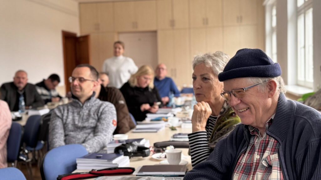 Starszy mężczyzna w granatowej czapce i kobieta obok niego siedzą przy stole i uśmiechają się, w tle inni uczestnicy przeglądają materiały