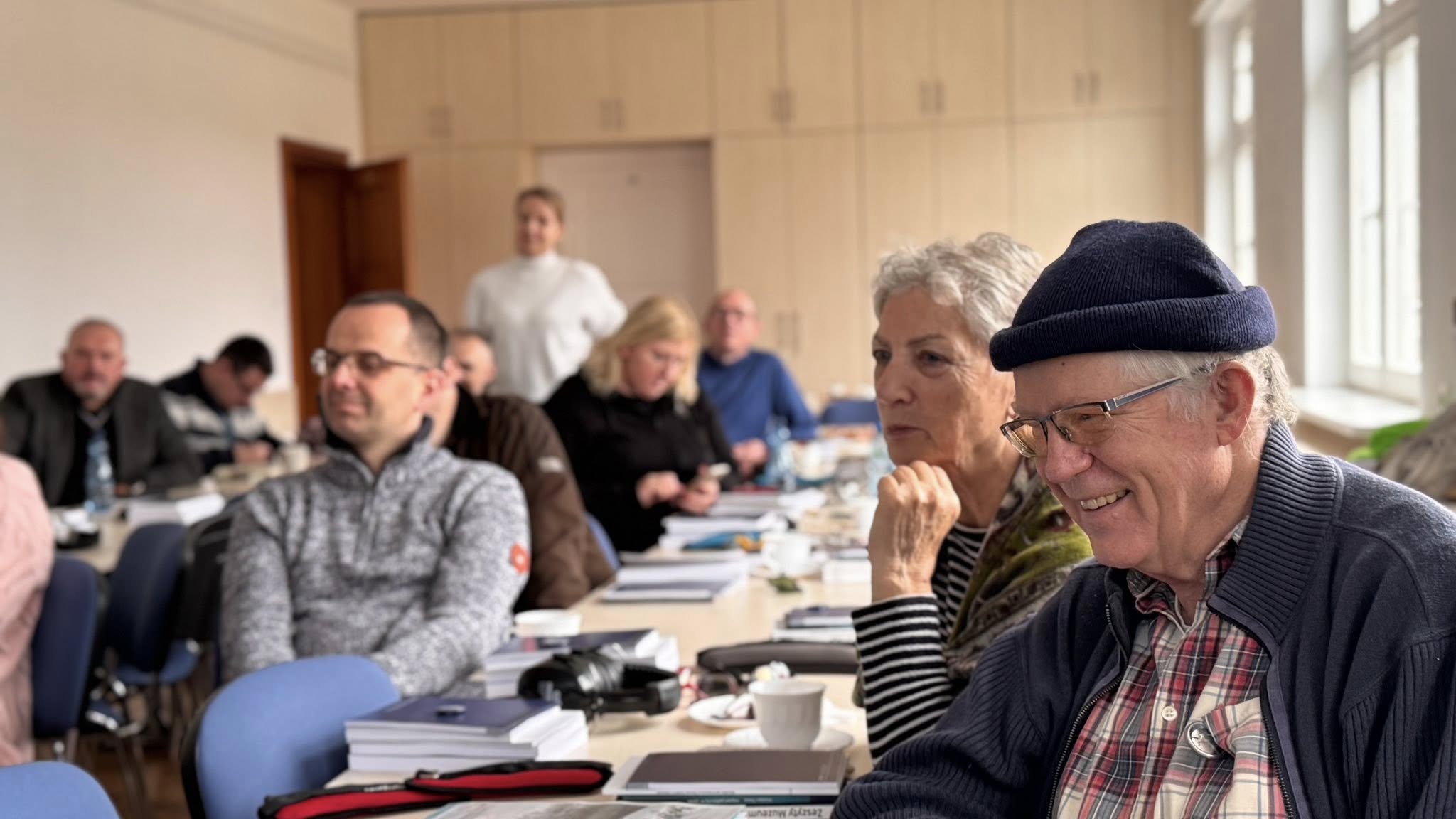 Starszy mężczyzna w granatowej czapce i kobieta obok niego siedzą przy stole i uśmiechają się, w tle inni uczestnicy przeglądają materiały