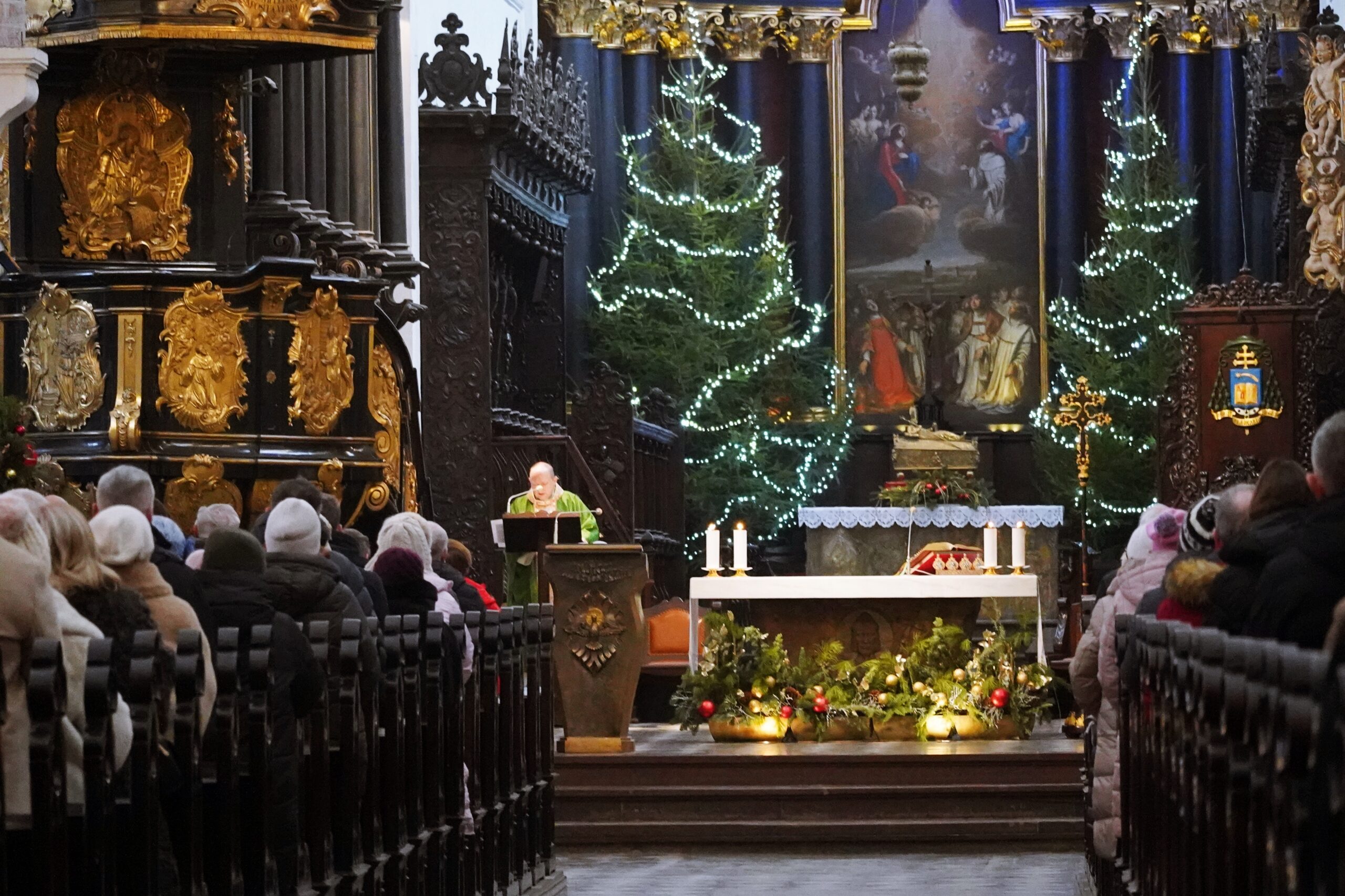 Widok na ołtarz w kościele podczas nabożeństwa; po bokach zgromadzeni wierni, a przy ambonie duchowny w zielonym ornacie. Ołtarz udekorowany, obok choinki z białymi światełkami.