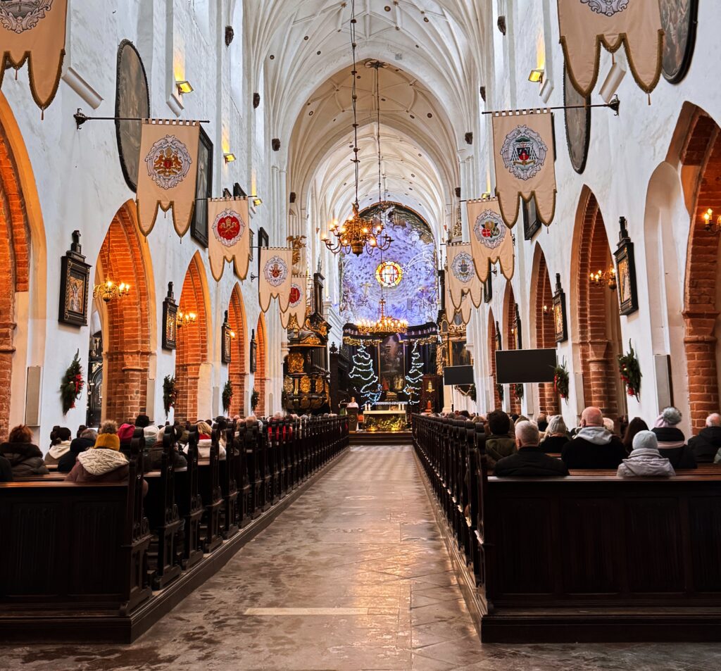 Szeroki kadr na wnętrze kościoła z długą nawą, ławkami i ludźmi siedzącymi po obu stronach przejścia; nad przejściem wiszą chorągwie, a w oddali widać ołtarz i dekoracje świąteczne.