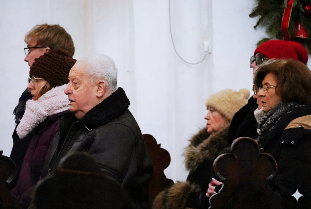 Zbliżenie na osoby stojące w kościele podczas nabożeństwa; na pierwszym planie kobieta w czapce i okularach oraz mężczyzna w ciemnej kurtce, w tle kolejne osoby i bożonarodzeniowa dekoracja.