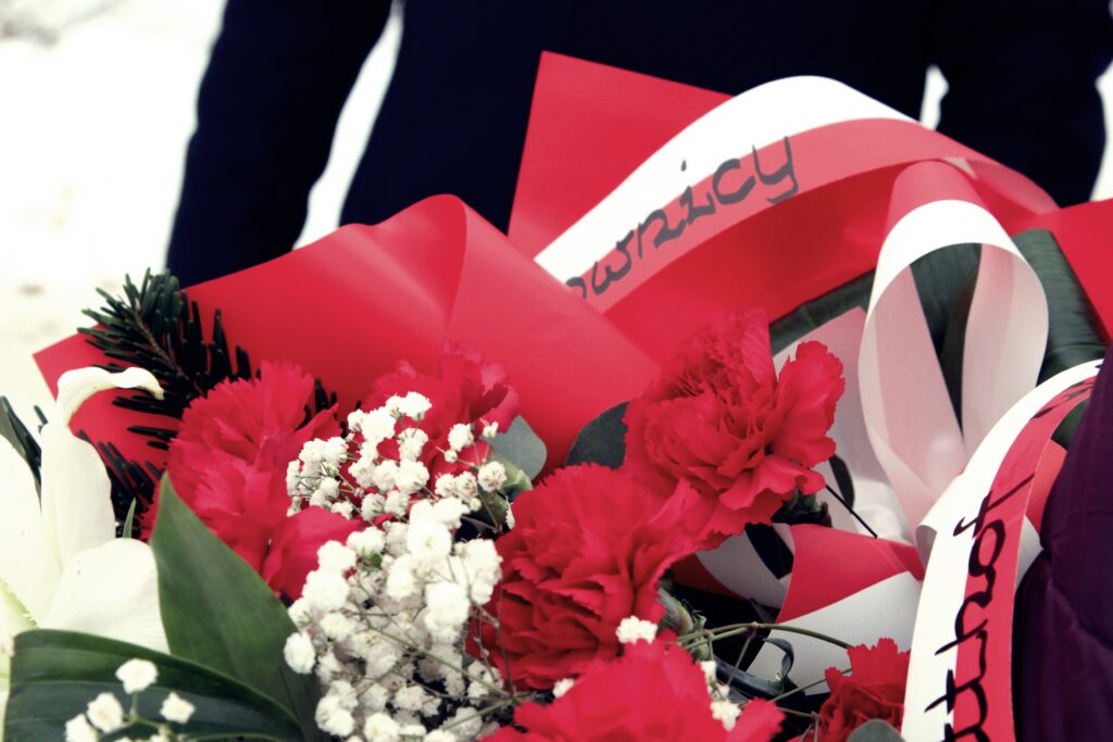 Zbliżenie na wiązankę z czerwonych goździków i białej gipsówki, owiniętą czerwonym papierem; widać biało-czerwoną wstęgę z napisem oraz iglaste gałązki.