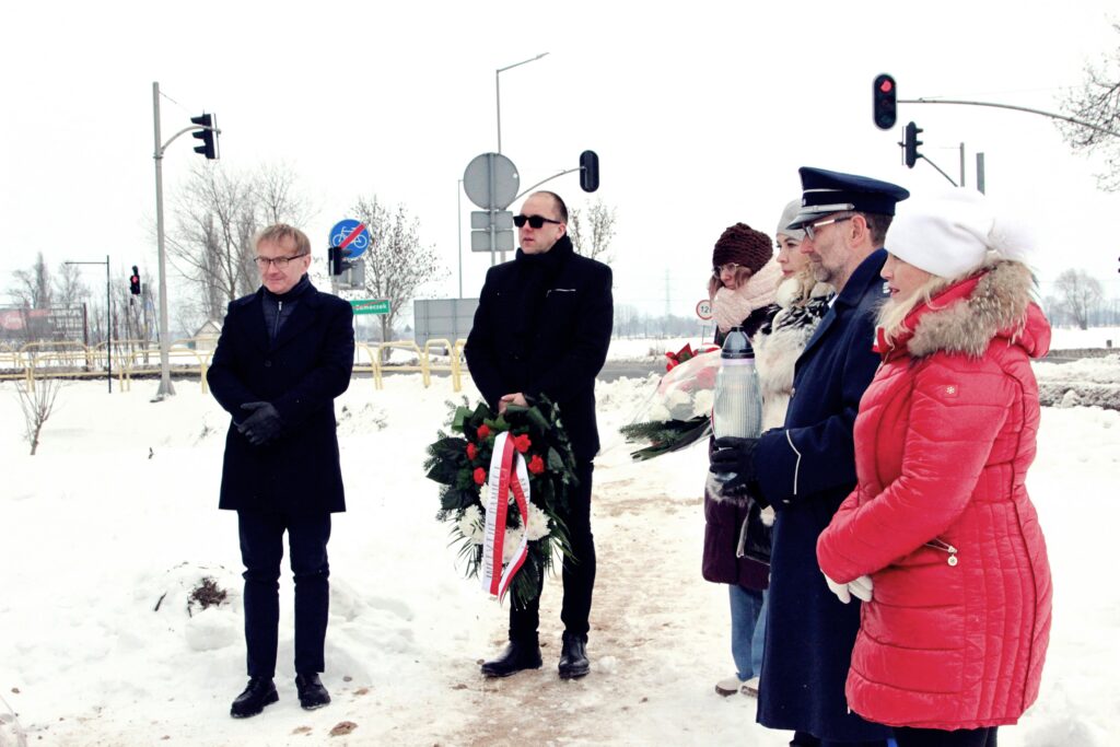 Trzy osoby stoją na śniegu obok tablicy informacyjnej; mężczyzna w mundurze i dwie kobiety, jedna trzyma znicz z biało-czerwoną wstążką; w tle zimowa zabudowa i baner gminy.