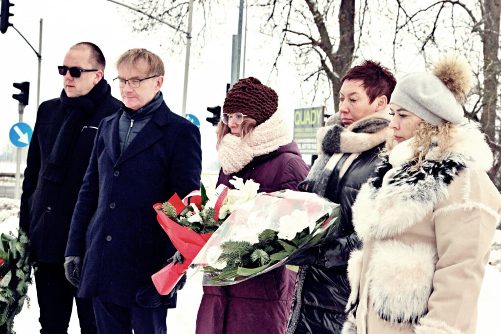 Pięć osób stoi w ciszy podczas uroczystości na śniegu; jedna osoba trzyma bukiet białych kwiatów w czerwonym papierze, w tle znaki drogowe i drzewa.