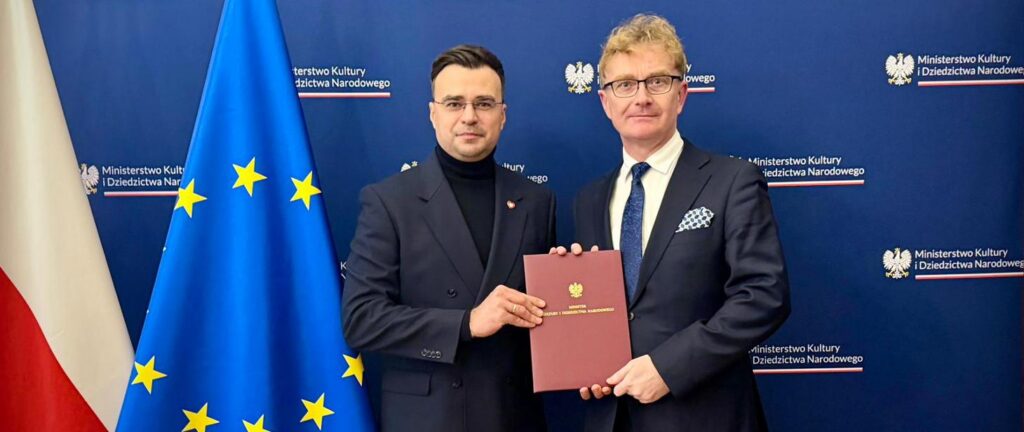 Dwóch mężczyzn w garniturach stoi na tle ścianki z logotypem Ministerstwa Kultury i Dziedzictwa Narodowego; wspólnie trzymają bordową teczkę z godłem. Po lewej widać flagę Polski i flagę Unii Europejskiej.