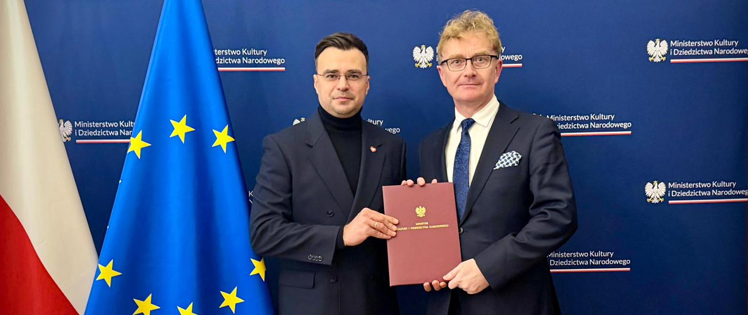 Dwóch mężczyzn w garniturach stoi na tle ścianki z logotypem Ministerstwa Kultury i Dziedzictwa Narodowego; wspólnie trzymają bordową teczkę z godłem. Po lewej widać flagę Polski i flagę Unii Europejskiej
