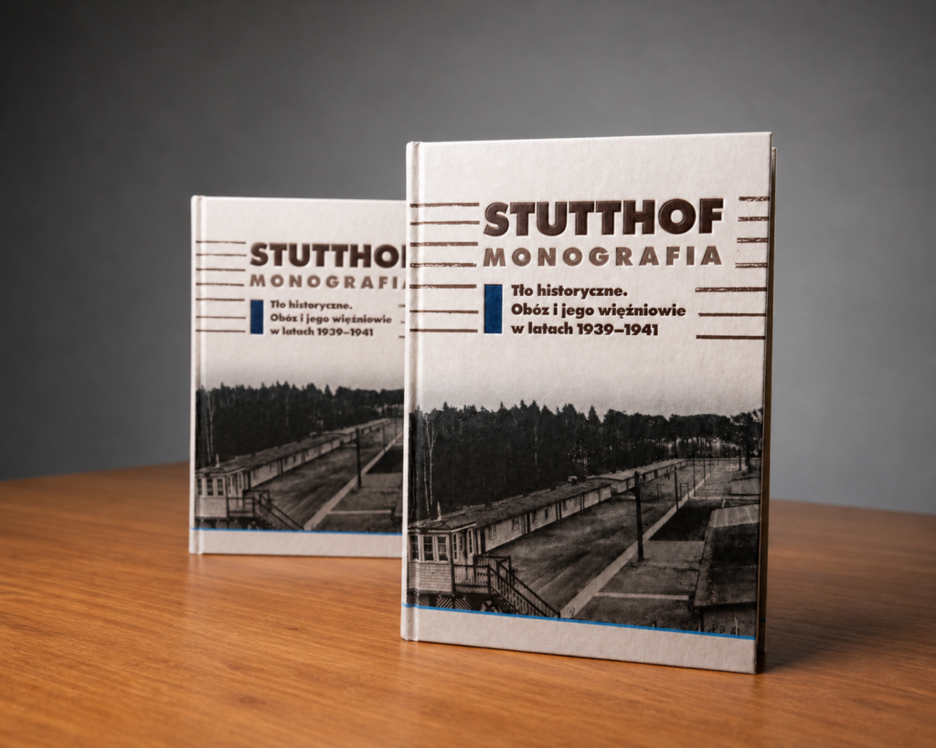 Fotografia przedstawia dwa stojące egzemplarze pierwszego tomu publikacji „Stutthof. Monografia. Tło historyczne. Obóz i jego więźniowie w latach 1939–1941”. Książki ustawiono na drewnianym blacie, jedna znajduje się bliżej obiektywu, druga lekko z tyłu i jest rozmyta. Na jasnej okładce widoczny jest duży tytuł „STUTTHOF. MONOGRAFIA” oraz czarno-biała fotografia przedstawiająca teren dawnego obozu z barakami i wieżyczką strażniczą. Tło zdjęcia jest neutralne, szare, a całość ma charakter studyjnej fotografii promującej publikację.