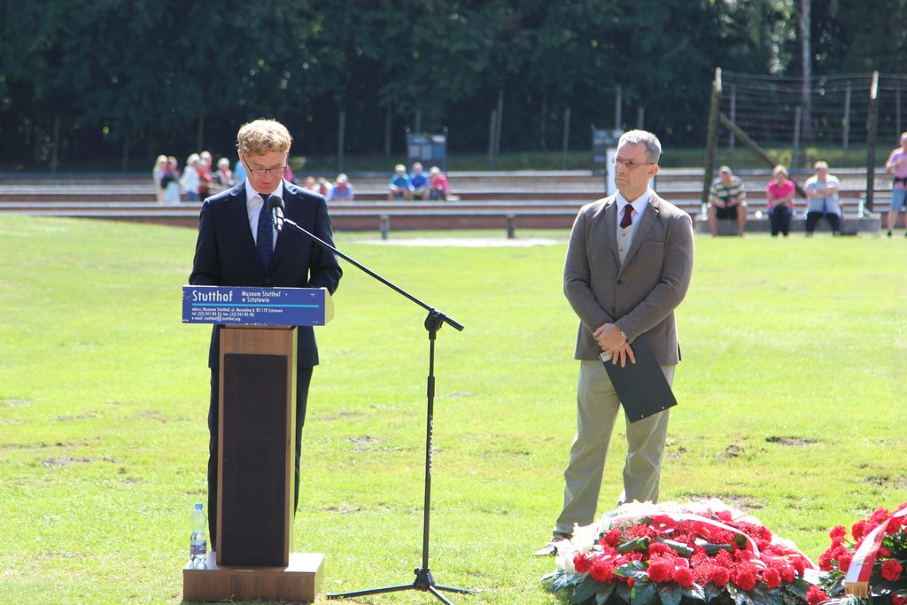 Przemówienie podczas uroczystości rocznicowej na terenie Muzeum Stutthof; przed mównicą leżą wieńce i kwiaty