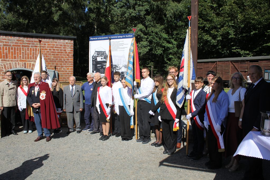 Uczestnicy uroczystości na terenie byłego KL Stutthof pozują do wspólnego zdjęcia. Wśród nich są poczty sztandarowe młodzieży oraz zaproszeni goście. W tle tablica „Pierwsze uroczystości na terenie byłego KL Stutthof”