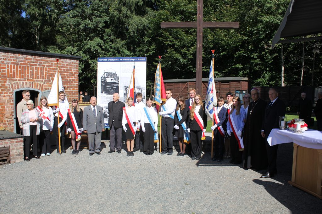 Grupowe zdjęcie uczestników uroczystości przy dużym drewnianym krzyżu na terenie byłego KL Stutthof. W centrum stoją poczty sztandarowe młodzieży, obok duchowni i pozostali uczestnicy.