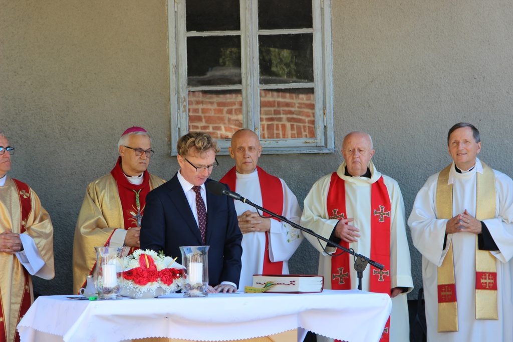 Mężczyzna w garniturze przemawia przy mikrofonie podczas uroczystości religijnej. Za ołtarzem stoją duchowni w szatach liturgicznych, wśród nich duchowny w mitrze biskupiej.