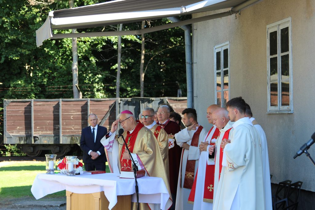 Duchowni podczas nabożeństwa przy polowym ołtarzu na terenie byłego KL Stutthof. Duchowny w mitrze biskupiej stoi przy mikrofonie i wykonuje gest błogosławieństwa.