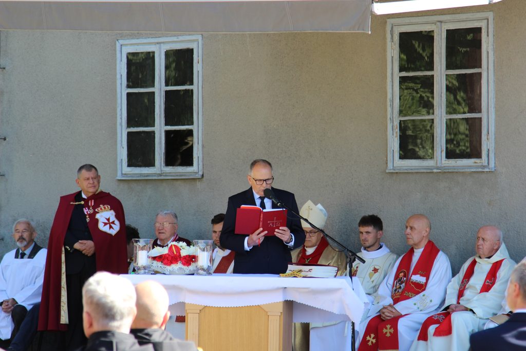 Mężczyzna w garniturze odczytuje tekst z czerwonej księgi przy mikrofonie. Za polowym ołtarzem siedzą duchowni uczestniczący w uroczystości.