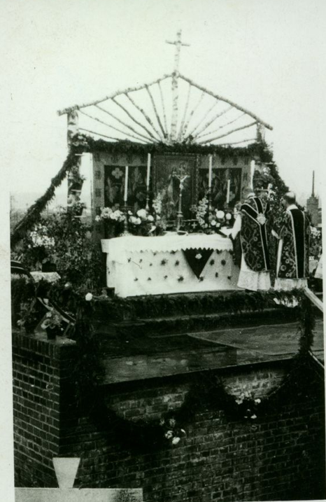 Czarno-biała fotografia przedstawiająca polowy ołtarz ustawiony na ceglanej konstrukcji. Przy udekorowanym kwiatami ołtarzu stoją dwaj duchowni w szatach liturgicznych. Nad ołtarzem znajduje się lekka konstrukcja zwieńczona krzyżem.