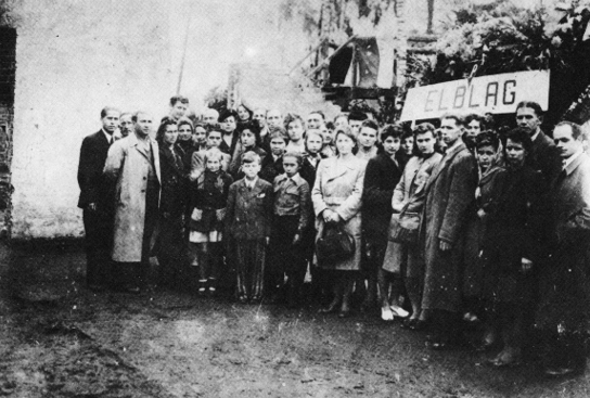 Czarno-biała fotografia grupowa przedstawiająca kilkadziesiąt osób, w tym kobiety, mężczyzn i dzieci, stojących na zewnątrz. Niektórzy mężczyźni mają na sobie pasiaste stroje więźniarskie. Po prawej stronie grupa trzyma tablicę z napisem „ELBLĄG”.