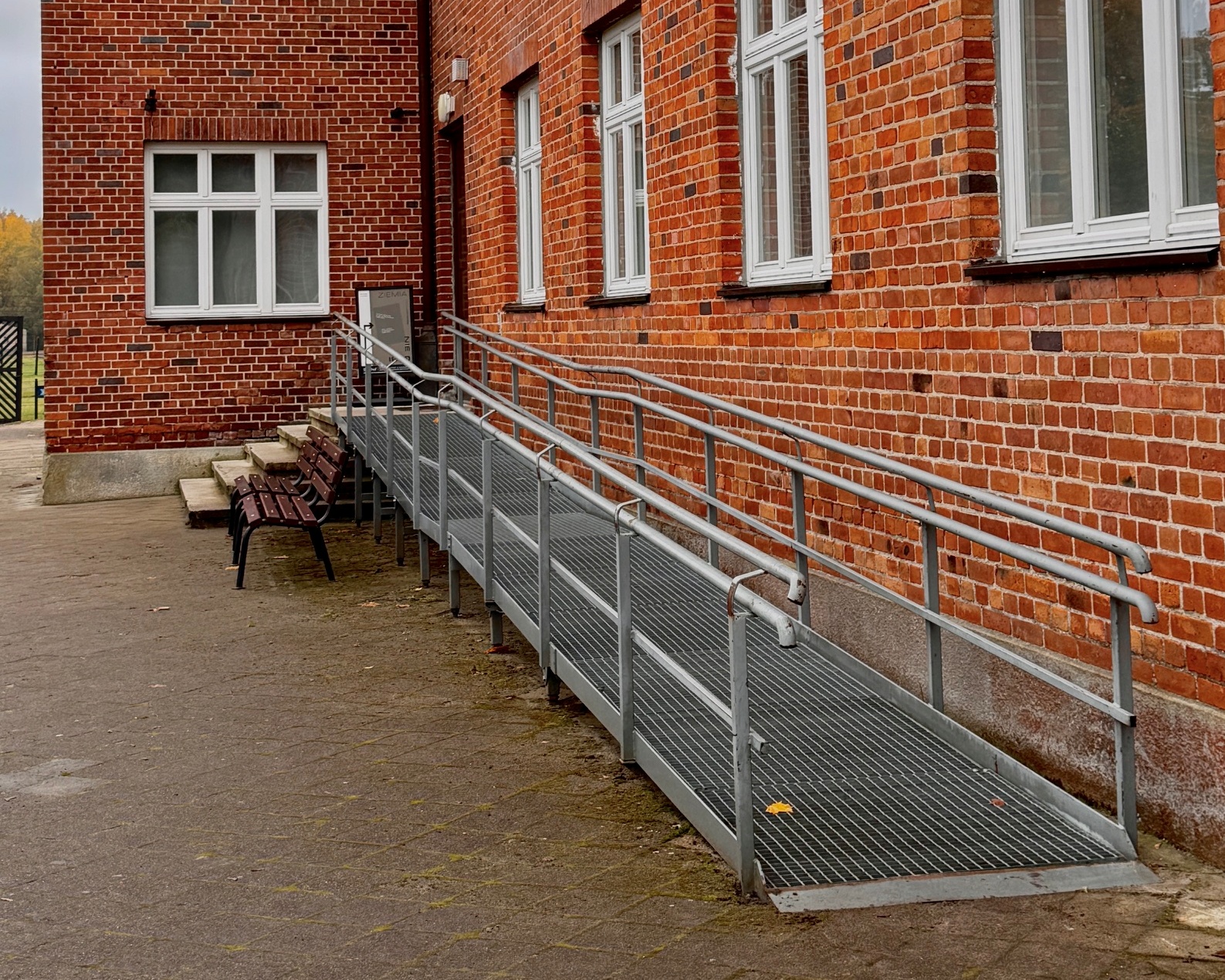 Metalowy podjazd dla osób poruszających się na wózkach, prowadzący do wejścia do ceglanego budynku administracyjnego Muzeum Stutthof w Sztutowie. Podjazd ma nawierzchnię z metalowej kraty i obustronne poręcze. Obok znajdują się schody oraz drewniane ławki.